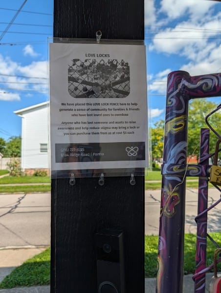 A laminated LOVE LOCKS sign on a purple-painted post, beside a chain-link gate hung with dozens of padlocks.